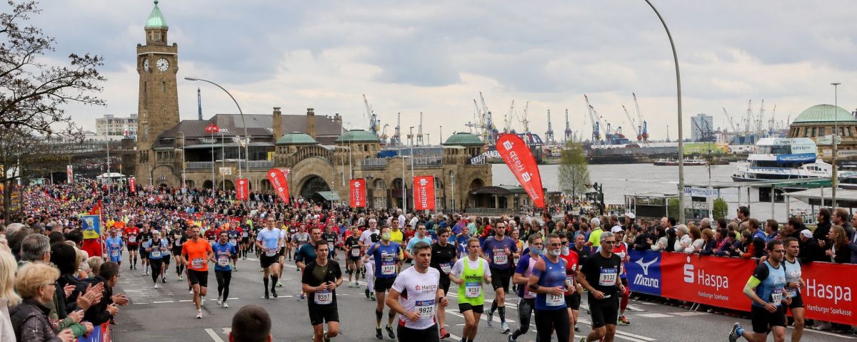 Läuferfeld beim Haspa Marathon Hamburg auf der Strecke entlang der Alster mit tausenden Zuschauern und Hamburger Wahrzeichen im Hintergrund.